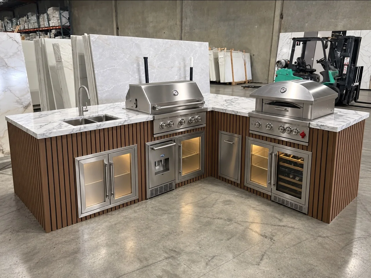 L-shaped outdoor kitchen island finished with teak and black slat cladding, with a grill, pizza oven, and sink